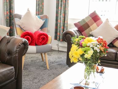 A living room with leather sofas and a patterned armchair with cushions and red rolled blankets a vase of flowers on a coffee table in Gwersyllt Benllech