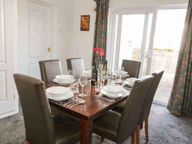 A dining room with a wooden table set for six with white dishes and wine bottles next to a sliding glass door with patterned curtains at Gwersyllt in Benllech