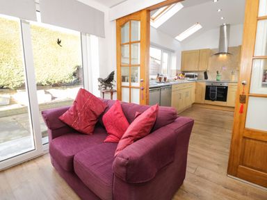 A kitchen with wooden cabinets and a purple sofa with red cushions in front of glass doors at Gwersyllt in Benllech