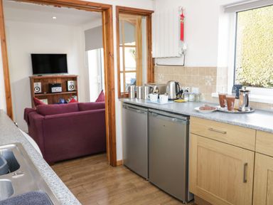 A kitchen with two small refrigerators and a countertop with mugs and a kettle next to a living room with a purple sofa and a television on a wooden stand at Gwersyllt in Benllech