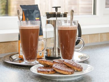 Two glasses of hot chocolate with spoons on a tray next to a French press and a packet of coffee with a plate of chocolate covered cookies on a kitchen counter at Gwersyllt in Benllech