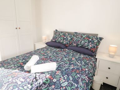 A bedroom with a bed with floral bedding two rolled white towels on the bed two white bedside tables with lamps a white wardrobe in the corner at Gwersyllt in Benllech