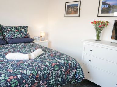 A bedroom with a floral bedspread a bedside table with a lamp a dresser with flowers in a vase and framed pictures on the wall at Gwersyllt in Benllech