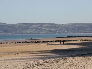 A sandy beach with scattered rocks and people walking near the shoreline with hills in the background at Gwersyllt in Benllech