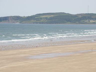 A sandy beach with a few people walking near the water and hills in the background at Gwersyllt in Benllech