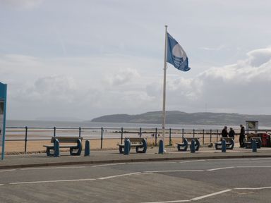 A seaside promenade with benches a flagpole and people near a sandy beach and ocean in Gwersyllt Benllech