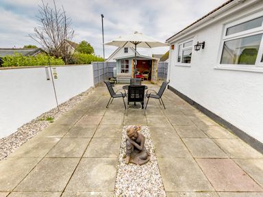 An outdoor patio area with a table and four chairs under an umbrella and a garden shed at the back with a statue in the center of the paved ground at Gwersyllt in Benllech