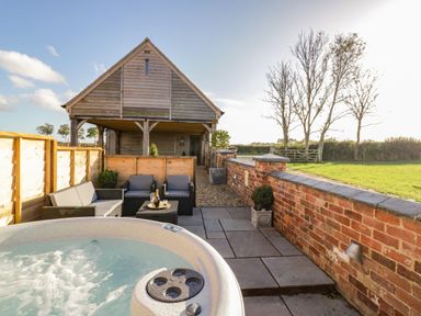 An outdoor patio with a hot tub seating area and a wooden building with a grassy field beyond at The Hyde in Armscote near Newbold on Stour