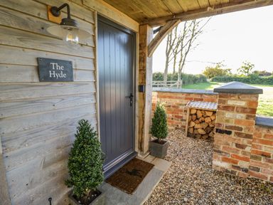 An outdoor entrance area with a gray door two potted plants a wall light and stacked firewood at The Hyde in Armscote near Newbold on Stour