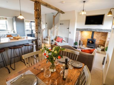 A dining table with plates and flowers next to a kitchen island with stools and a living area with a fireplace and television at The Hyde in Armscote near Newbold on Stour