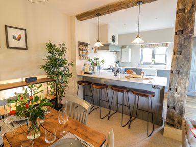A kitchen with a breakfast bar and stools next to a dining table with flowers and place settings at The Hyde in Armscote near Newbold on Stour