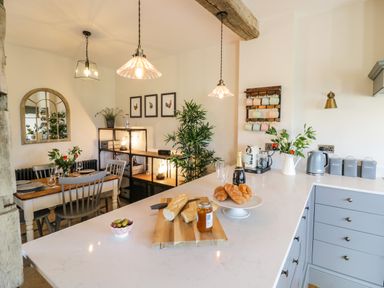A kitchen and dining area with a wooden dining table set and a kitchen island with croissants and bread at The Hyde in Armscote near Newbold on Stour