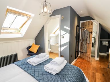 A bedroom with a bed with folded towels a chair with a cushion a wooden door with a robe hanging and an open doorway leading to a bathroom at The Hyde in Armscote near Newbold on Stour