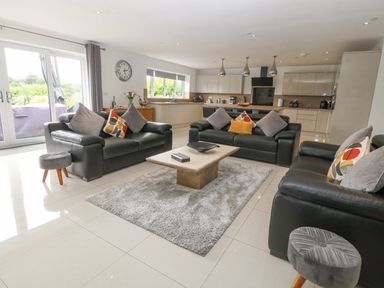 A living room with three black leather sofas and a coffee table on a rug with a kitchen in the background at Berllan in Llanbedrgoch near Benllech