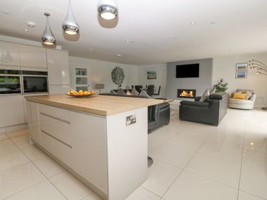 A modern open plan kitchen and living room with an island, black sofa, wall-mounted TV, fireplace, and dining table at Berllan in Llanbedrgoch near Benllech