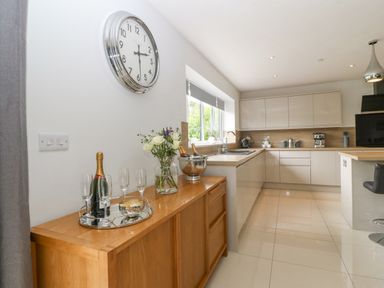 A kitchen with a wooden sideboard holding champagne and glasses a wall clock and white cabinets at Berllan in Llanbedrgoch near Benllech