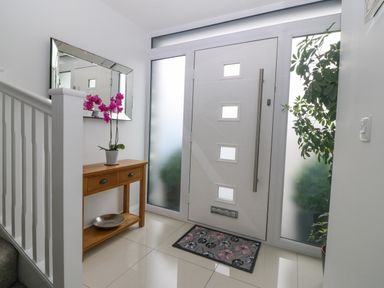 A hallway with a wooden console table with a pink orchid plant a large mirror tiled floor a gray staircase and a white front door with frosted glass windows at Berllan in Llanbedrgoch near Benllech