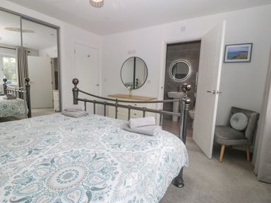 A bedroom with a metal bed frame and patterned bedspread two rolled towels on the bed a dresser with a round mirror a gray chair and an open door leading to a bathroom at Berllan in Llanbedrgoch near Benllech