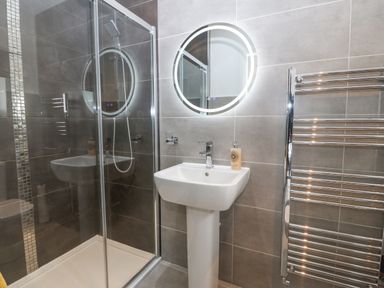 A bathroom with a shower enclosure a pedestal sink a round illuminated mirror and a heated towel rail at Berllan in Llanbedrgoch near Benllech