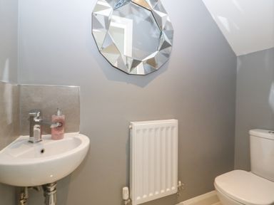 A bathroom with a white sink and soap dispenser a wall-mounted radiator a geometric mirror and a white toilet at Berllan in Llanbedrgoch near Benllech