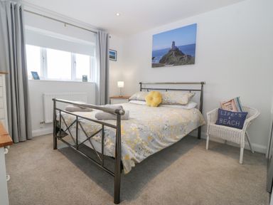 A bedroom with a metal bed frame bed a basket chair with cushions a bedside table with lamp and a window with curtains at Berllan in Llanbedrgoch near Benllech