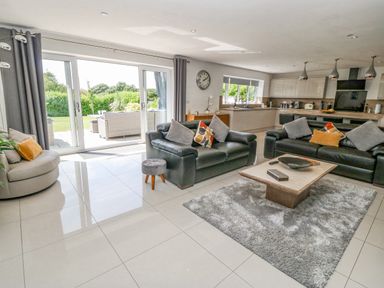An open living room and kitchen area with black sofas a round chair a coffee table on a rug and large sliding glass doors at Berllan in Llanbedrgoch near Benllech