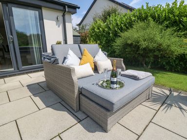 An outdoor patio with a wicker sofa with cushions and pillows and a tray with a bottle and glasses at Berllan in Llanbedrgoch near Benllech