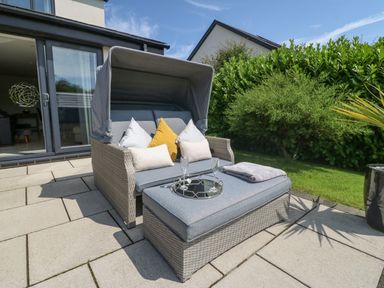 An outdoor patio with a wicker loveseat with cushions and a matching ottoman with a tray holding two glasses at Berllan in Llanbedrgoch near Benllech