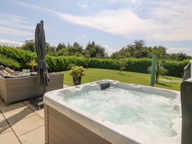 An outdoor patio with a hot tub a seating area with sofas an umbrella and a grassy garden with trees at Berllan in Llanbedrgoch near Benllech