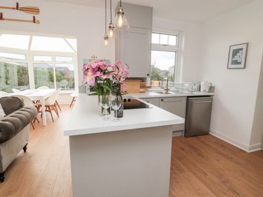 A kitchen with a white island countertop holding pink flowers two wine glasses and a bottle next to a gray cabinet with a stove a window a sink dishwasher and a dining area with a table and chairs visible at Shileen in Seahouses