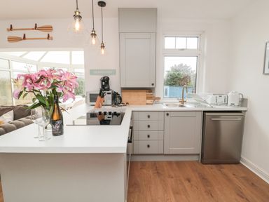 A kitchen with grey cabinets a white countertop wooden floor and a bottle of wine with glasses at Shileen in Seahouses