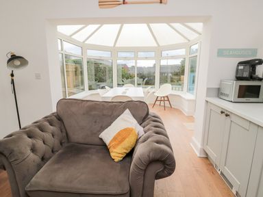 A living room with a brown sofa and a windowed dining area with a white table and chairs at Shileen in Seahouses