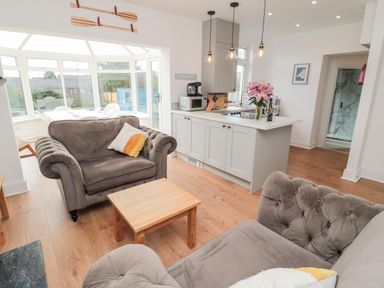A living room with gray armchairs a wooden coffee table and an open kitchen with white cabinets and hanging lights at Shileen in Seahouses