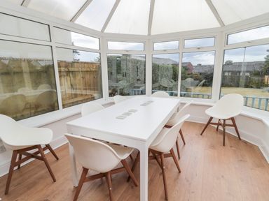 A dining area with a white table and six white chairs with wooden legs in a conservatory with windows overlooking a garden at Shileen in Seahouses