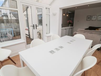 A dining area with a white table and six white chairs near glass doors and windows at Shileen in Seahouses