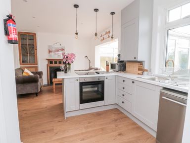 A kitchen with white cabinets an oven and a sink next to a living area with sofas and a fireplace at Shileen in Seahouses