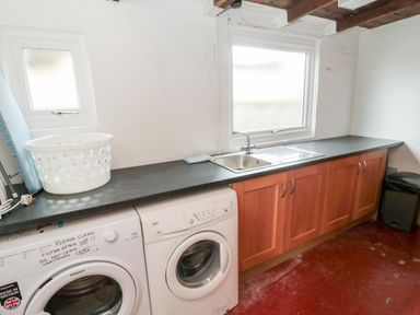A utility room with a washing machine, dryer, sink, countertop, ironing board, laundry basket, and wooden cabinets at Shileen in Seahouses