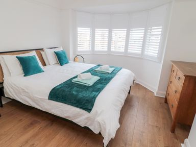 A bedroom with a bed with white and teal bedding and teal pillows next to a wooden dresser and a curved window with shutters at Shileen in Seahouses