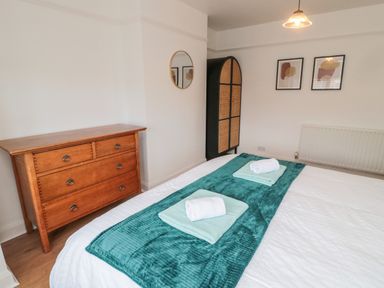A bedroom with a wooden dresser a bed with towels and a turquoise runner a black and rattan wardrobe and two framed artworks on the wall at Shileen in Seahouses