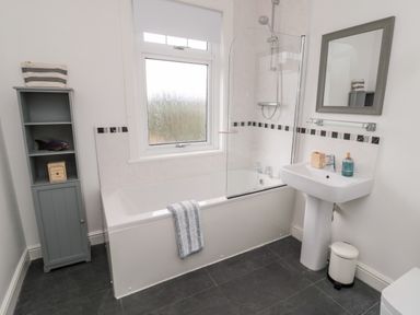 A bathroom with a bathtub shower combination a sink with a mirror above and a gray storage cabinet at Shileen in Seahouses