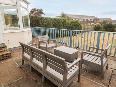 An outdoor patio with gray plastic chairs a bench and a table next to a blue railing and a yard at Shileen in Seahouses