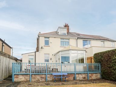 The exterior of a two-story semi detached house with a wooden deck and blue railing in a garden at Shileen in Seahouses