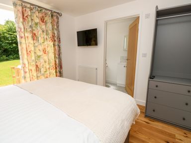 A bedroom with a bed facing a floral curtain window a wall mounted television an open door leading to a bathroom and a gray wardrobe with drawers at Barolo in Seaview