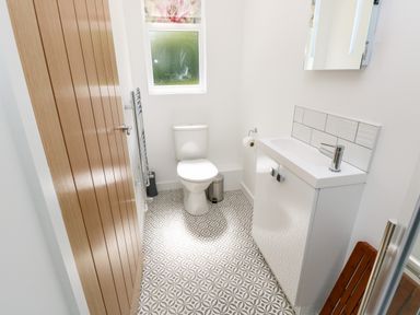 A bathroom with a toilet, a sink with cabinet, a mirror, a wooden door, a window with a floral blind, and patterned floor tiles at Cava in Seaview