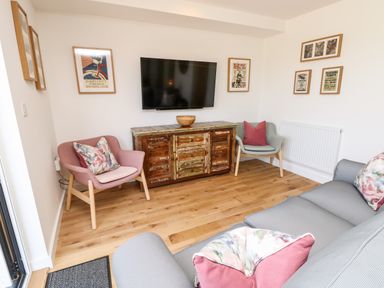 A sitting room with a wall-mounted TV a wooden cabinet two chairs a grey sofa and framed pictures on white walls at Claret in Seaview