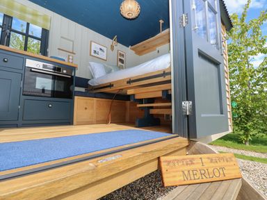 A small interior space with a bed, blue cabinets, an oven, wooden storage, and an open door showing greenery outside at Merlot in Pondwell near Seaview