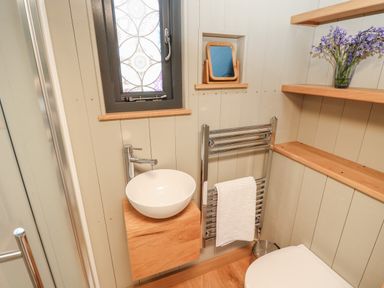 A small bathroom with a round white sink on a wooden cabinet a towel rail with a white towel flowers on a wooden shelf and a window with patterned glass at Merlot Pondwell near Seaview