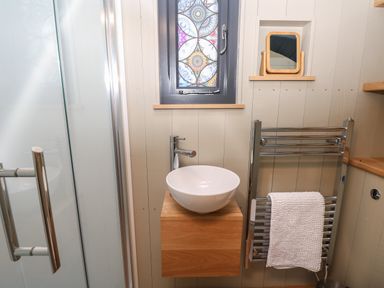 A small bathroom with a shower, a round white sink on a wooden cabinet, a towel rack with a white towel, a small window with patterned glass, and a small wooden-framed mirror Malbec Pondwell near Seaview