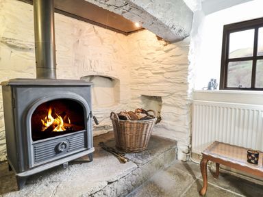 A wood burning stove with fire in a stone room next to a basket of firewood and a wooden table at Tan Y Garth Cottage in Glyndyfrdwy near Llangollen