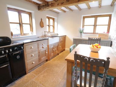 A kitchen with wooden cabinets a farmhouse sink a wooden dining table with chairs and a bowl of fruit at Tan Y Garth Cottage in Glyndyfrdwy near Llangollen